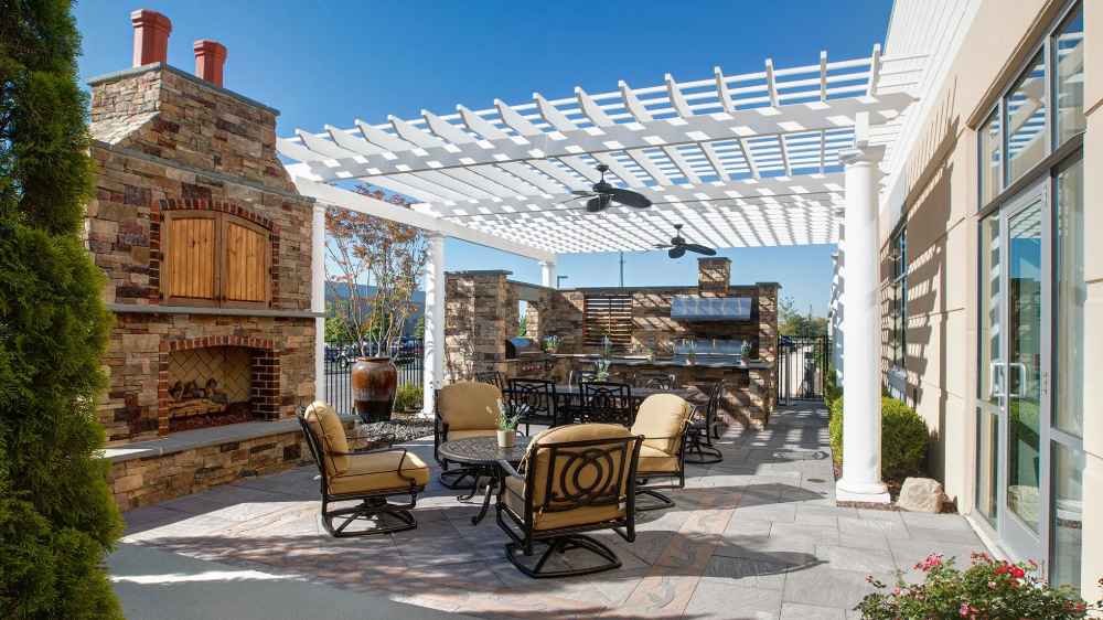 A stone fireplace and outdoor kitchen sit under a white Structureworks Trex Pergola with ceiling fans and elegant patio furniture