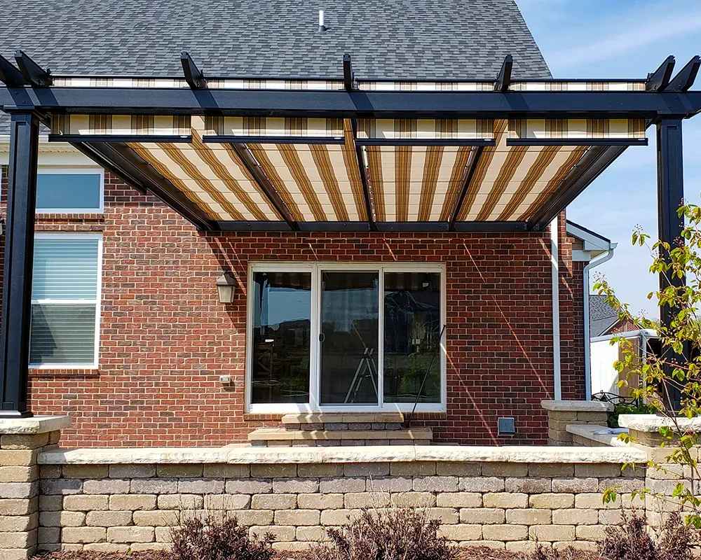 A striped fabric-roof Structureworks Atlas Aluminum Pergola extends from a brick house over a small patio with a low stone wall