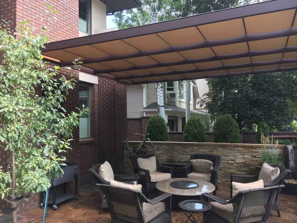 A stylish patio setup with wicker chairs surrounds a fire table under a wide, taupe-hued Structureworks Craftsman Aluminum Patio Cover & Pergola against a brick home