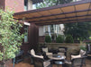 A stylish patio setup with wicker chairs surrounds a fire table under a wide, taupe-hued Structureworks Craftsman Aluminum Patio Cover & Pergola against a brick home