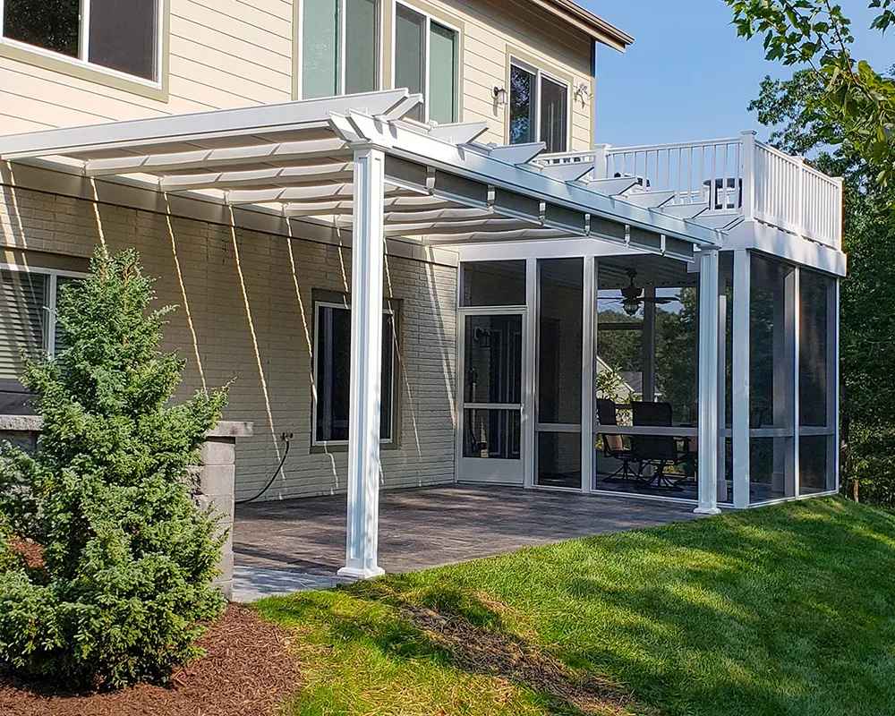 A white Structureworks Atlas Aluminum Pergola is attached to a two-story house, shading a patio with glass screen walls below a balcony