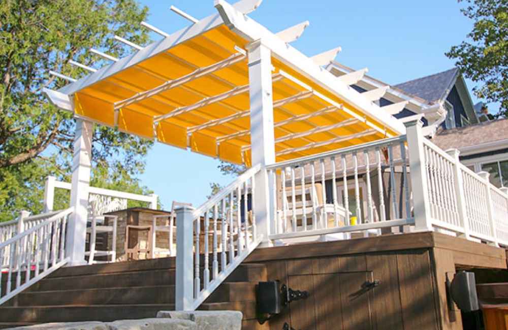 A white Structureworks Atlas Aluminum Pergola with a vibrant yellow retractable canopy shades a deck area attached to a home with white railings