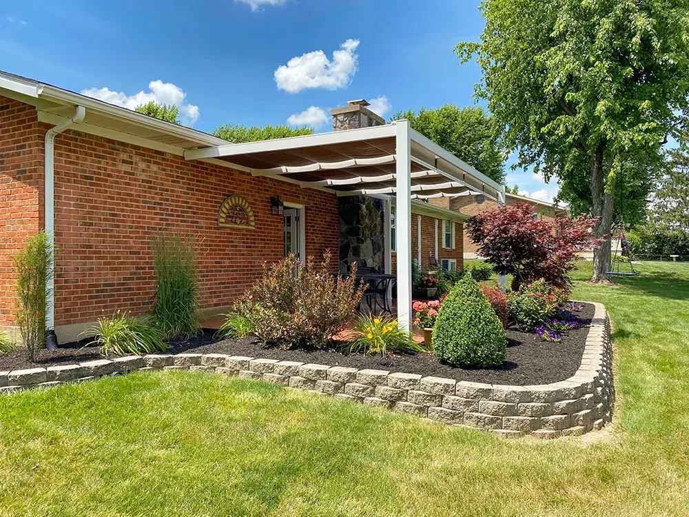A white Structureworks Outlook Aluminum Patio Cover & Pergola overhangs a flower-lined patio on a red-brick home, viewed from a side angle with green lawn in front