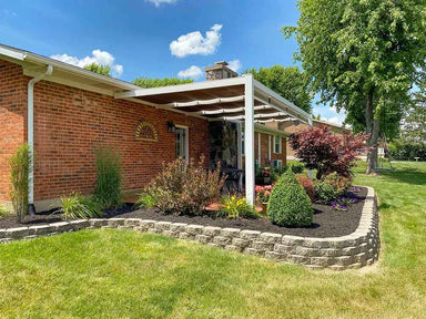 A white Structureworks Outlook Aluminum Patio Cover & Pergola overhangs a flower-lined patio on a red-brick home, viewed from a side angle with green lawn in front