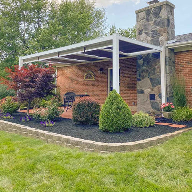 A white Structureworks Outlook Aluminum Patio Cover & Pergola shelters a red-brick patio with black furniture and lush landscaping