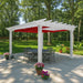 A white free-standing Structureworks Atlas Aluminum Pergola with a bright red canopy shades a round patio table in a wooded backyard