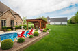 A beautiful backyard features a Brookside Timber Frame Glazier Hideaway Pavilion by the poolside, surrounded by lush greenery and colorful Adirondack chairs