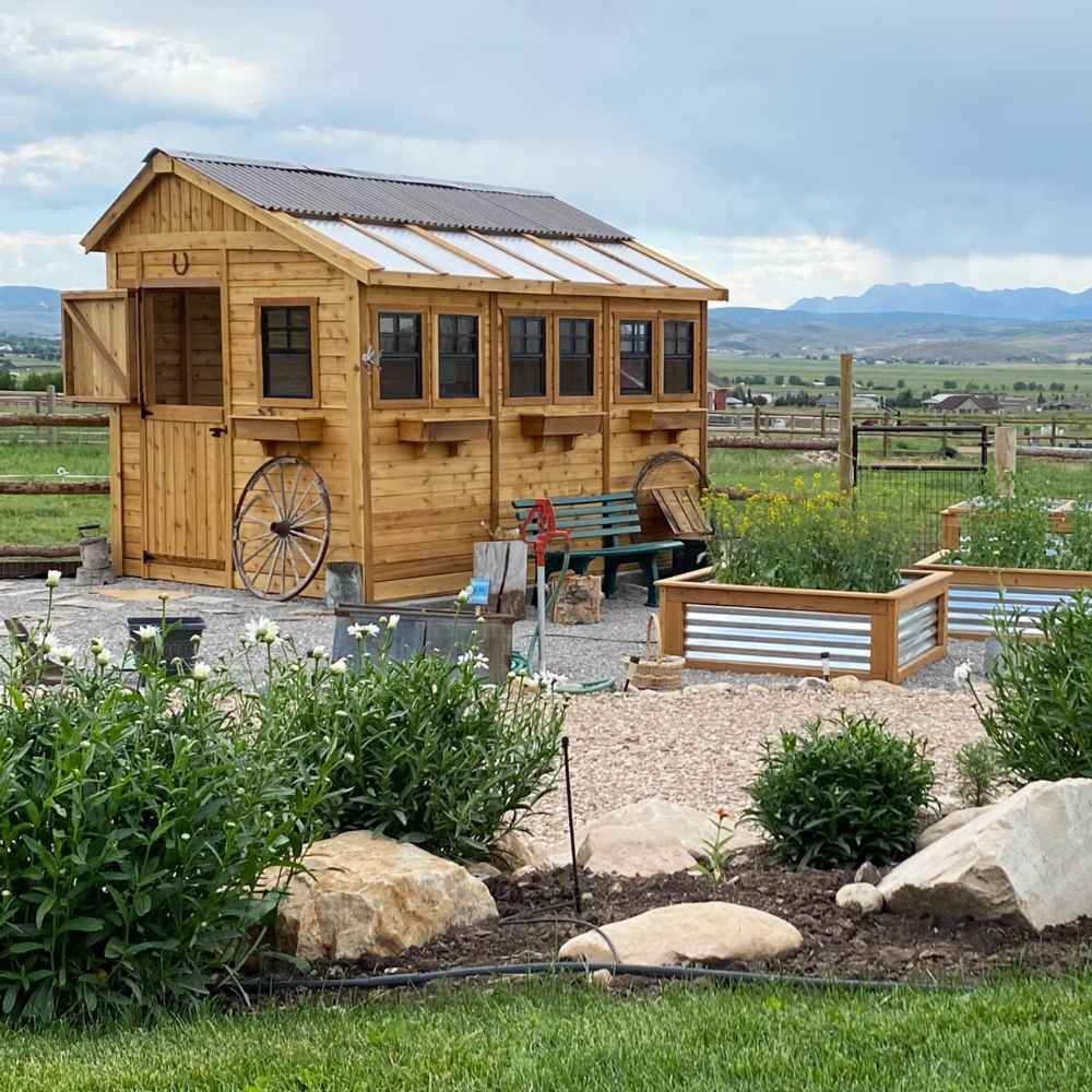 A charming garden scene shows the Outdoor Living Today 8×12 Sunshed Garden Shed with a wooden wagon wheel and raised garden beds nearby under cloudy skies