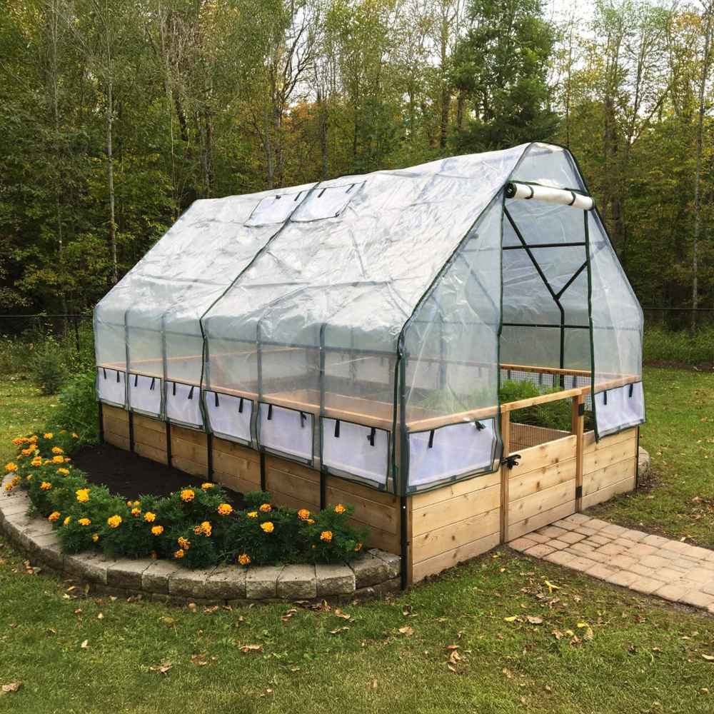A clear plastic greenhouse cover stretches over an Outdoor Living Today 8×16 Raised Garden Bed, surrounded by grass and a flower bed with bright orange blooms