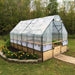 A clear plastic greenhouse cover stretches over an Outdoor Living Today 8×16 Raised Garden Bed, surrounded by grass and a flower bed with bright orange blooms