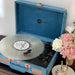 A close-up of an Arkrocket Curiosity Suitcase Bluetooth Turntable (Ocean Velvet) with a transparent vinyl record spinning placed beside pink and white flowers