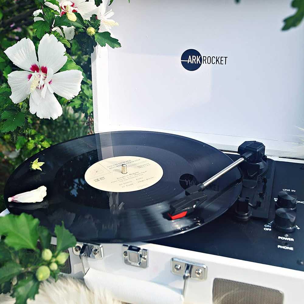 A close-up of a spinning vinyl record on Arkrocket Curiosity Suitcase Bluetooth Turntable (White) placed outdoors near vibrant white flowers