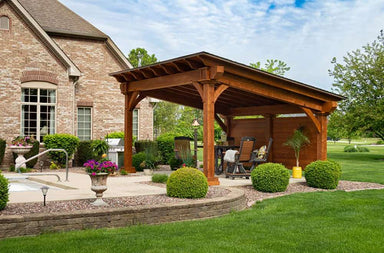 A close-up of the Brookside Timber Frame Glazier Hideaway Pavilion showcases its sturdy wooden structure, cozy seating area, and well-manicured plants