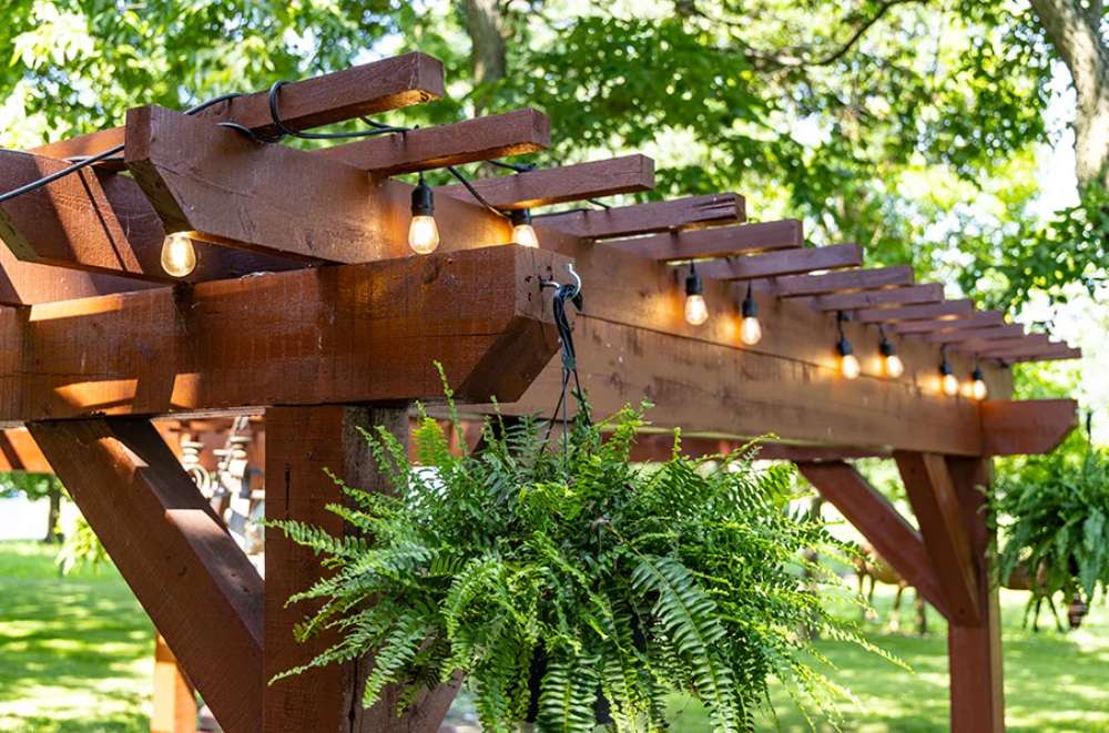 A close-up of the Brookside Timber Frame Shadelan Retreat Pergola highlights its sturdy wooden beams, glowing string lights, and lush hanging ferns