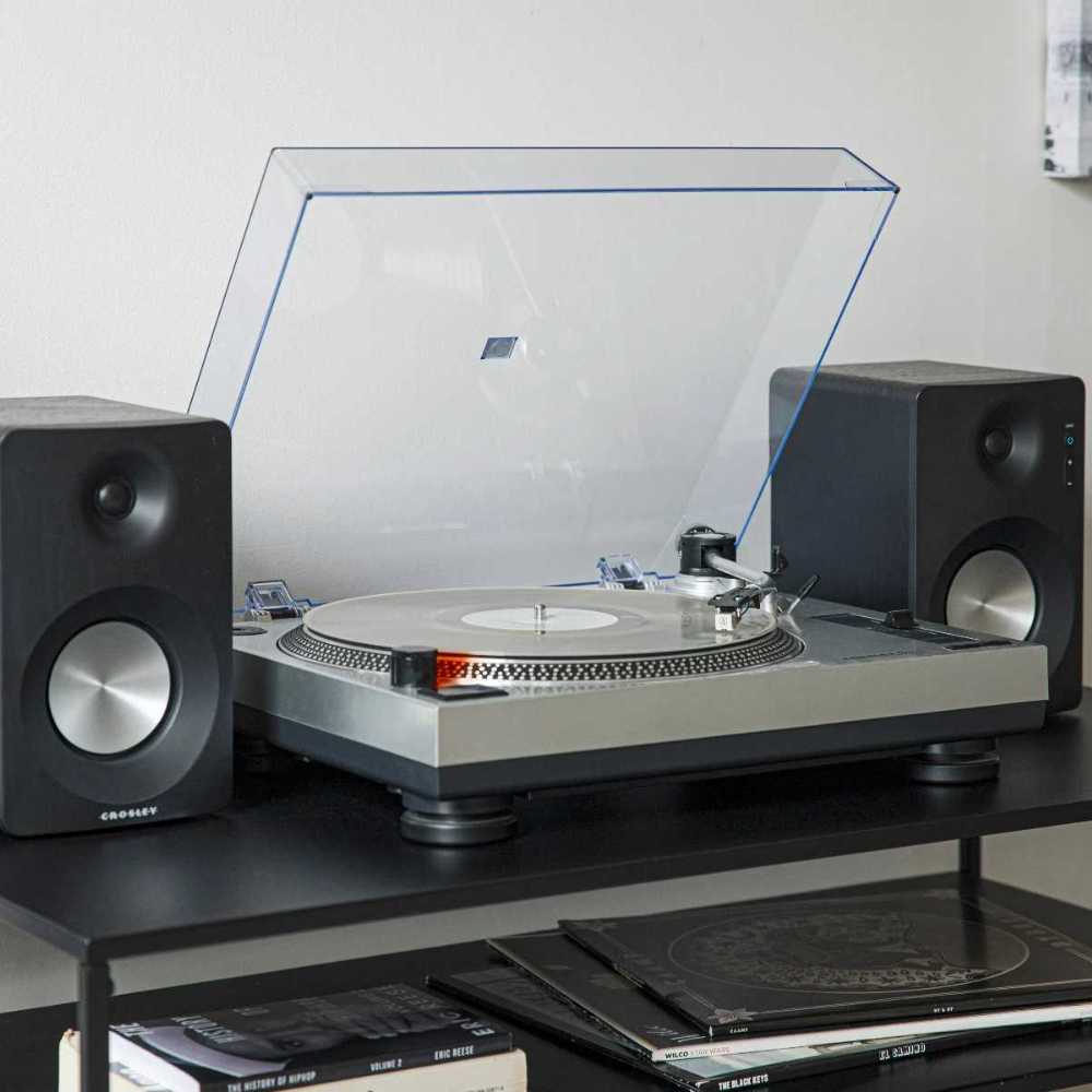 A close-up of the Crosley K100A Record Player with S100 Speakers - Silver highlights its clear dust cover open while the turntable plays a record