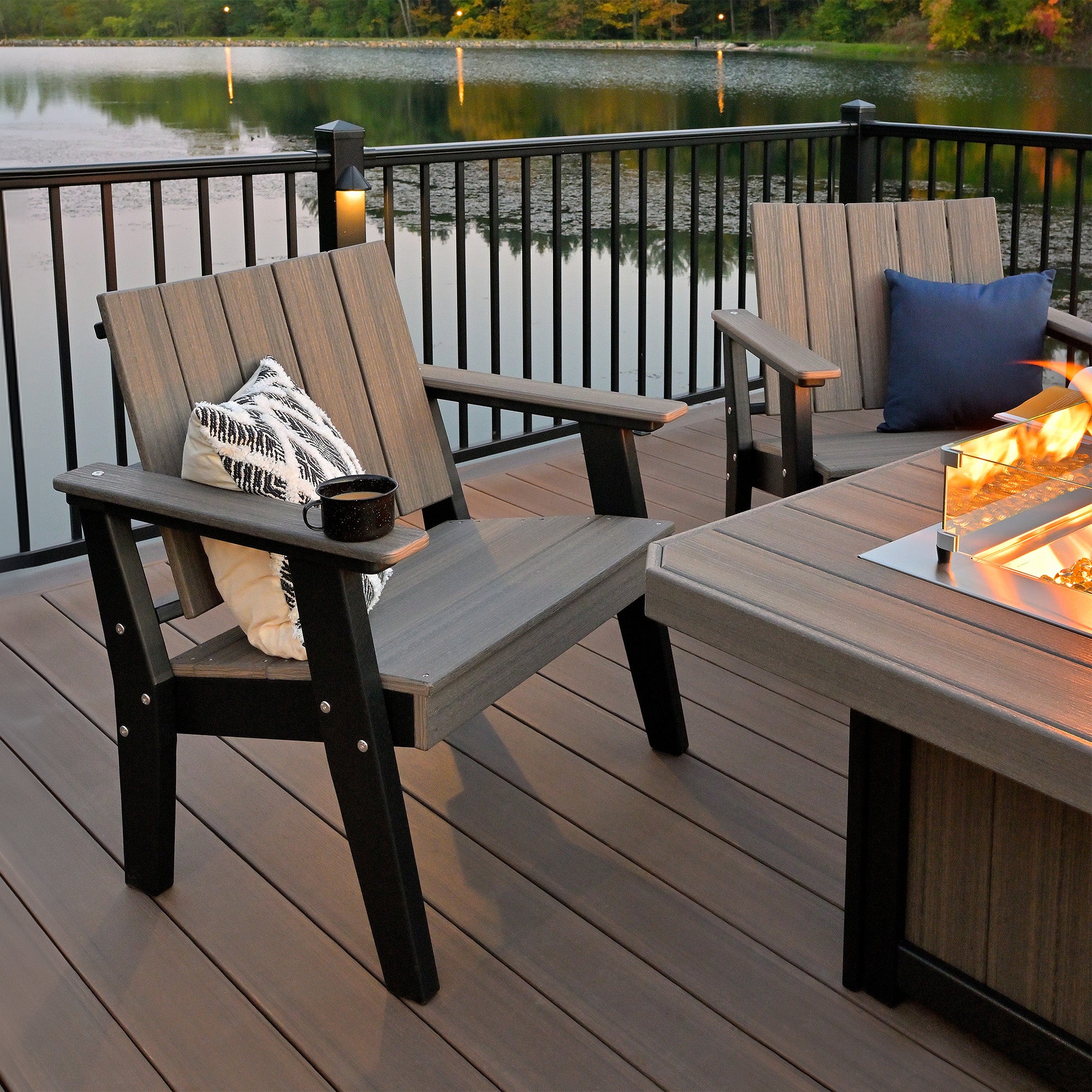 A close-up of the LuxCraft Hearthside Furniture Set highlights a sturdy Adirondack chair with a stylish cushion and a warm beverage resting on the armrest, set against a peaceful lakeside view