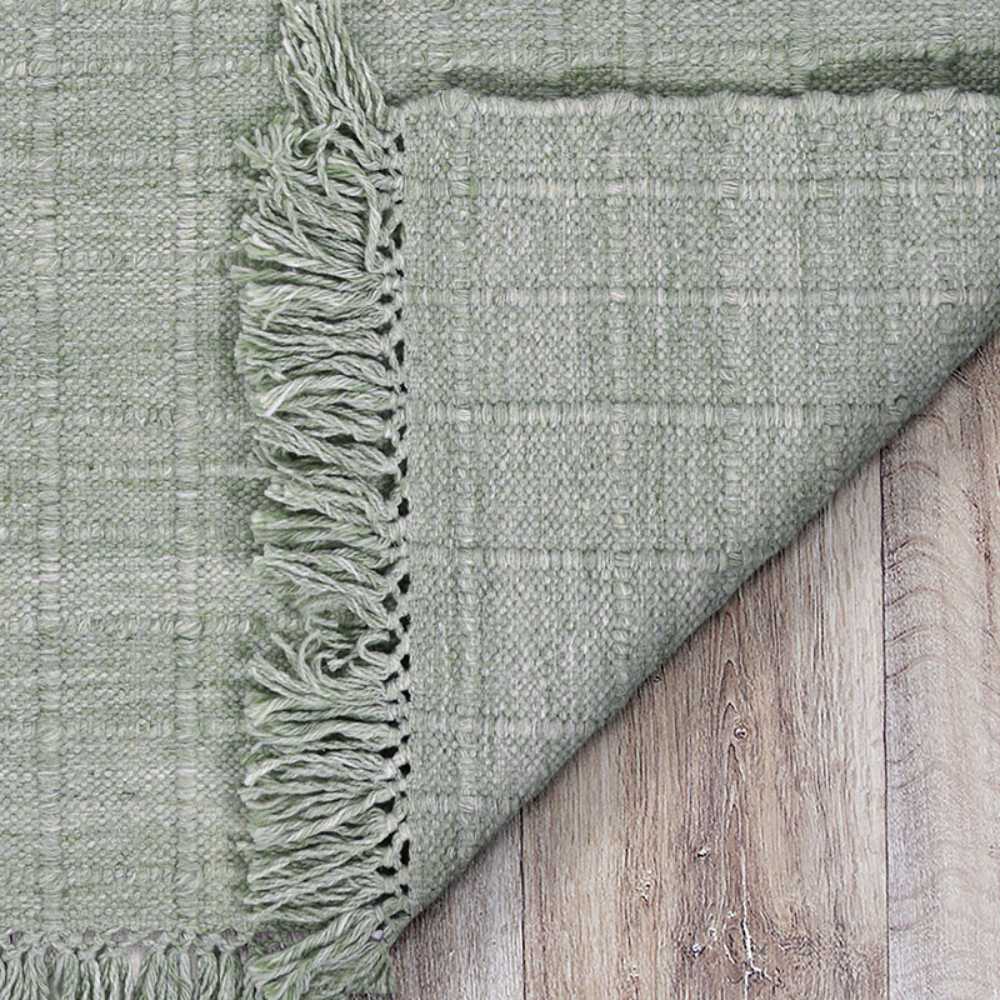 A close-up of the corner reveals the Couristan Ascot Indoor/Outdoor Rug - Windowpane/Celedon flipped over on wooden flooring, highlighting its woven texture and tassels
