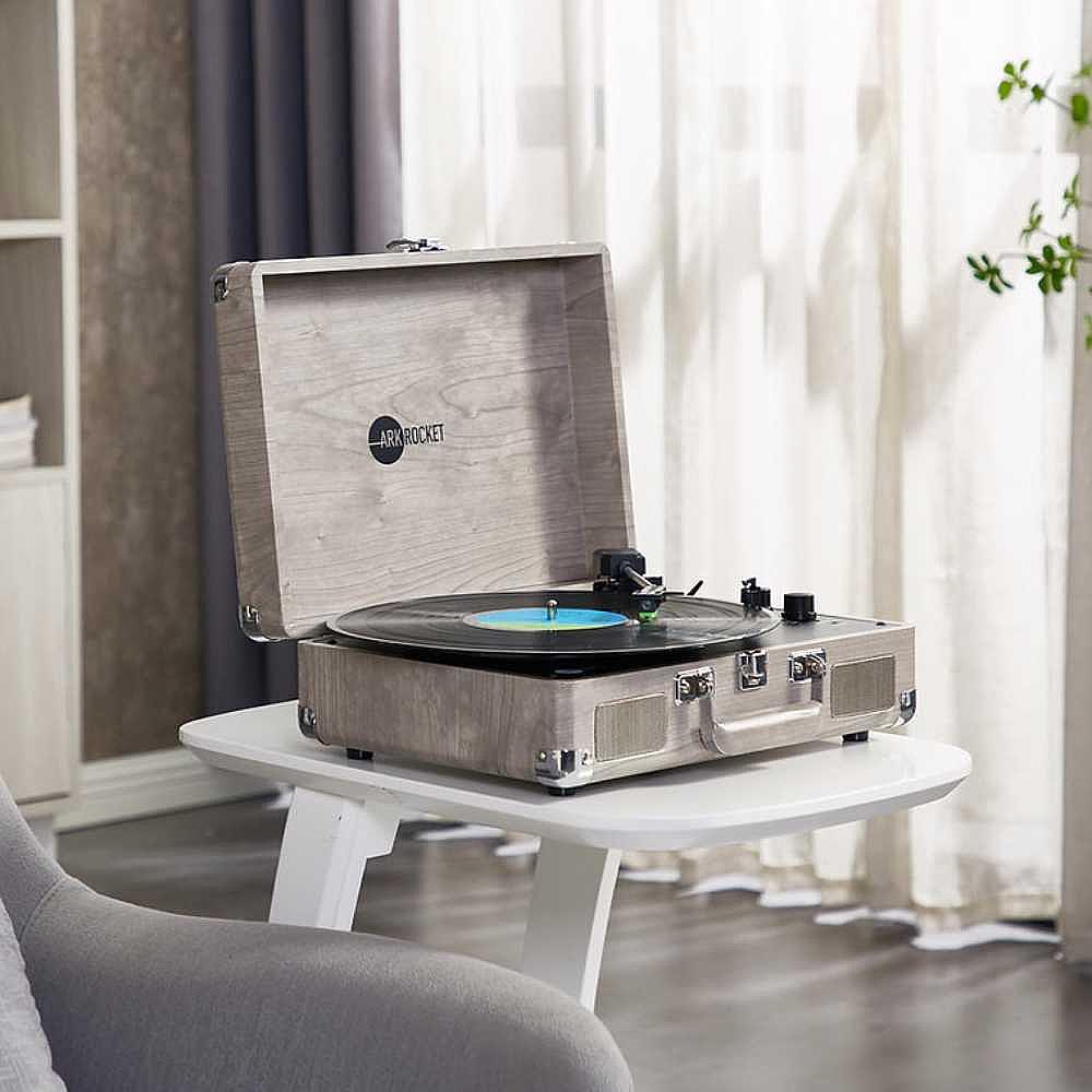 A close-up shot shows Arkrocket Curiosity Suitcase Bluetooth Turntable (Gray Wood Grain) on a small white table with a vinyl record playing