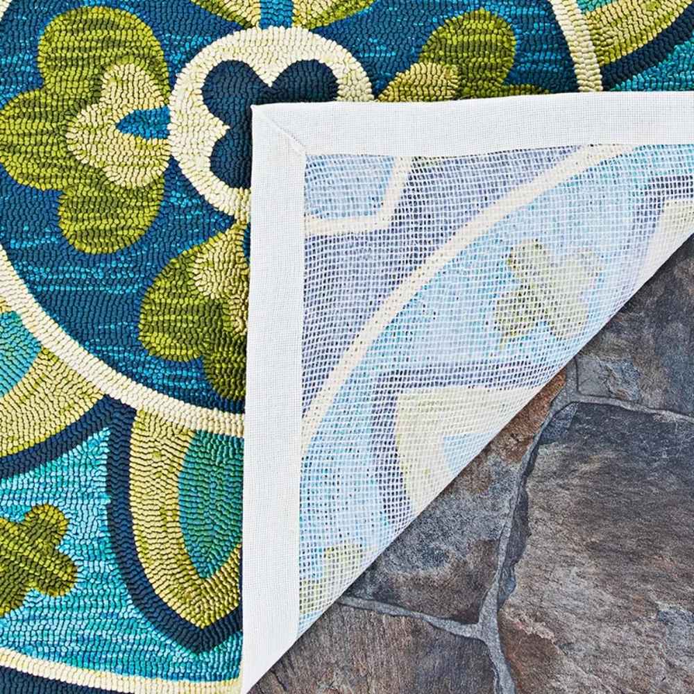 A close-up shows the back corner of the Couristan Covington Indoor/Outdoor Rug - Rip Tide/Ocean-Green lifted to reveal its mesh backing on a stone surface