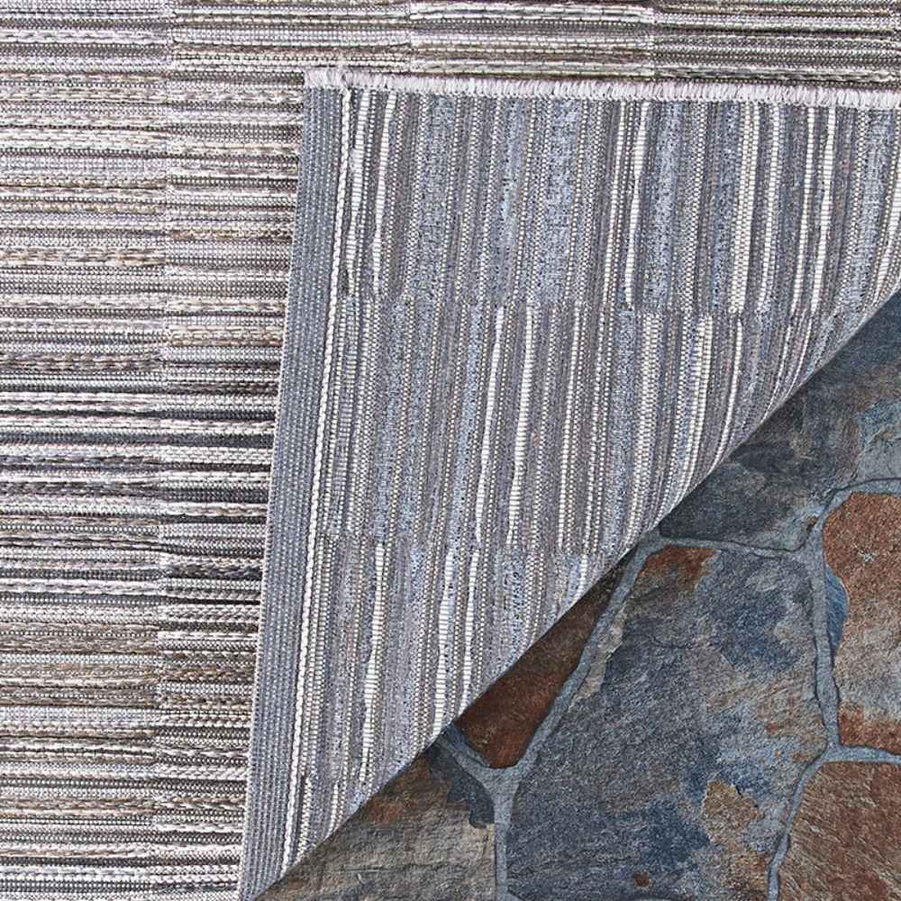 A close-up shows the corner of the Couristan Cape Indoor/Outdoor Rug - Fayston/Silver-Charcoal flipped up to reveal its detailed texture and backing on stone flooring