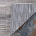 A close-up shows the corner of the Couristan Cape Indoor/Outdoor Rug - Fayston/Silver-Charcoal flipped up to reveal its detailed texture and backing on stone flooring