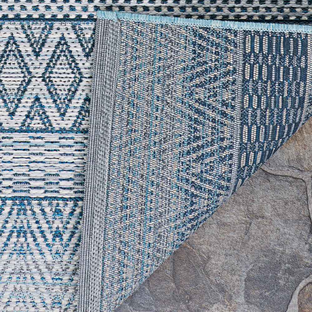 A close-up shows the corner of the Couristan Cape Indoor/Outdoor Rug - Gables/Surf flipped over, revealing its detailed blue and ivory woven patterns and finished edges