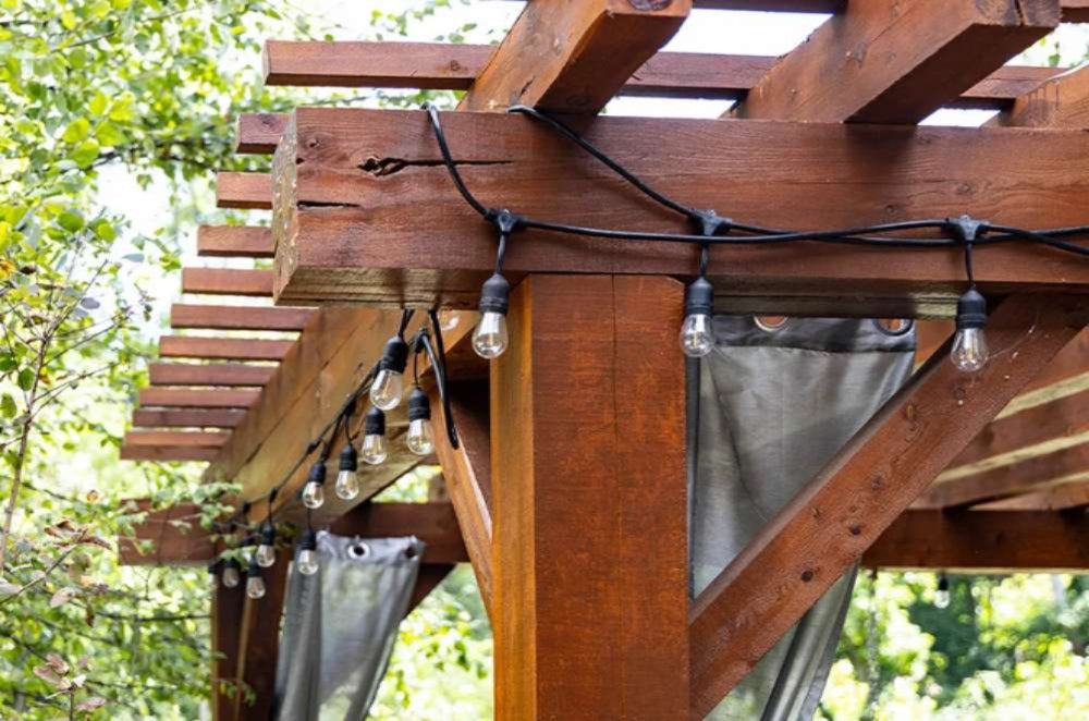 A close-up view of the Brookside Timber Frame Highlan Oasis Pergola showcases its sturdy wooden beams, elegantly draped curtains, and charming hanging string lights