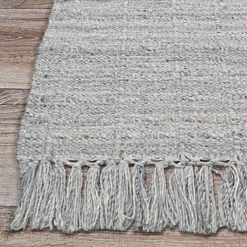 A corner of the Couristan Ascot Indoor/Outdoor Rug - Windowpane/Grey reveals its thick weave and evenly spaced tassels on light wooden flooring