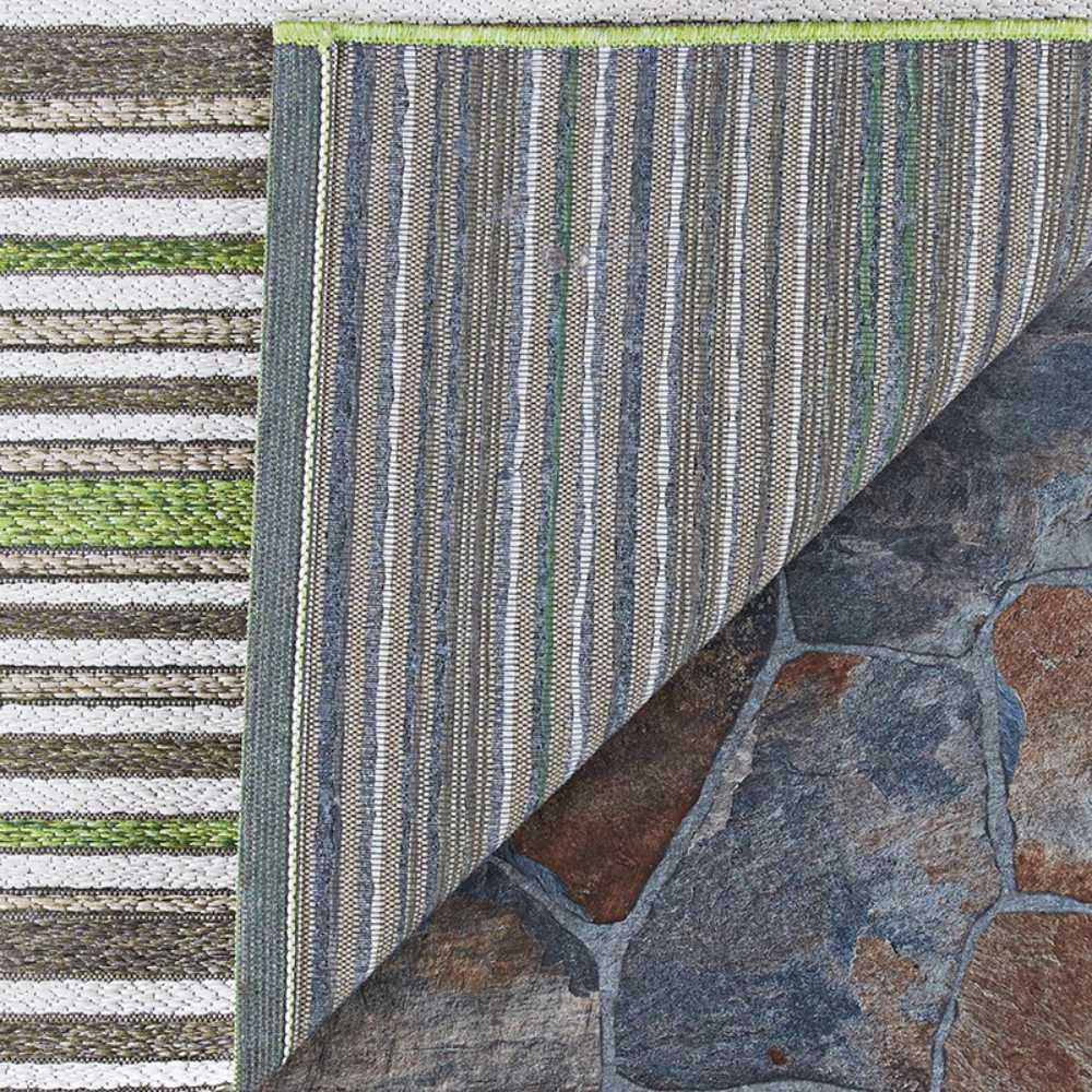 A corner of the Couristan Cape Indoor/Outdoor Rug - Brockton/Hunter Green-Brown is lifted to show its striped backing against a stone floor