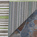 A corner of the Couristan Cape Indoor/Outdoor Rug - Brockton/Hunter Green-Brown is lifted to show its striped backing against a stone floor
