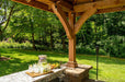 A cozy and inviting outdoor bar area under the Brookside Timber Frame Forest Melody Pavilion, complete with a stone countertop, refreshing drinks, and a view of the lush lawn