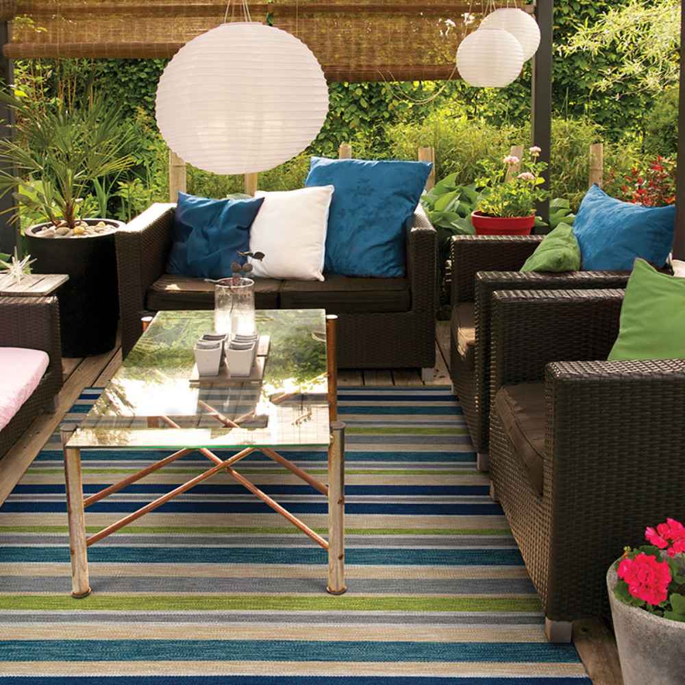 A cozy outdoor patio setup features the Couristan Cottages Indoor/Outdoor Rug - Alki/Ocean Shapes under a glass coffee table and wicker seating, surrounded by lush greenery