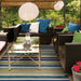 A cozy outdoor patio setup features the Couristan Cottages Indoor/Outdoor Rug - Alki/Ocean Shapes under a glass coffee table and wicker seating, surrounded by lush greenery