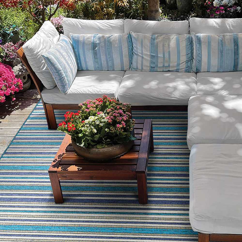 A cozy outdoor seating area with white cushioned sofas and a wooden table features the Couristan Cape Indoor/Outdoor Rug - Brockton/Cobalt-Teal adding vibrant striped color beneath