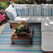 A cozy outdoor seating area with white cushioned sofas and a wooden table features the Couristan Cape Indoor/Outdoor Rug - Brockton/Cobalt-Teal adding vibrant striped color beneath