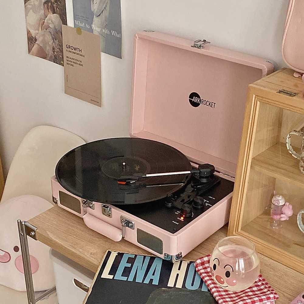 A cozy room features the Arkrocket Curiosity Suitcase Bluetooth Turntable (Pale Pink) on a side table playing a vinyl record next to decor