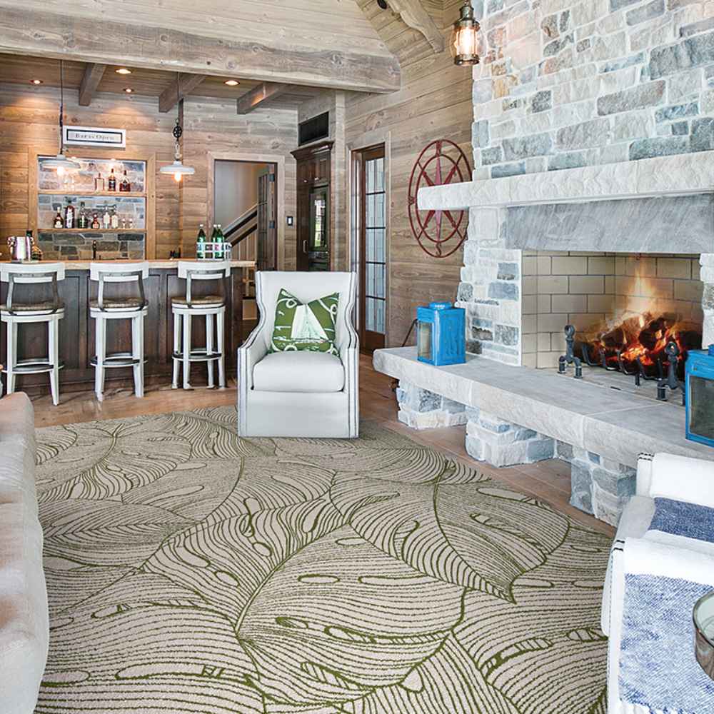 A cozy rustic living room features a stone fireplace and a large leafy-patterned Couristan Castille Indoor/Outdoor Rug - Palma/Petrol covering the wooden floor