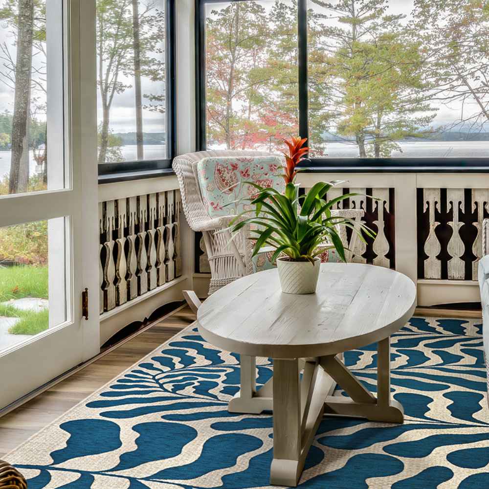 A cozy sunroom overlooks a lake, with a white coffee table and chair sitting on the Couristan Castille Indoor/Outdoor Rug - Fiore/Petrol