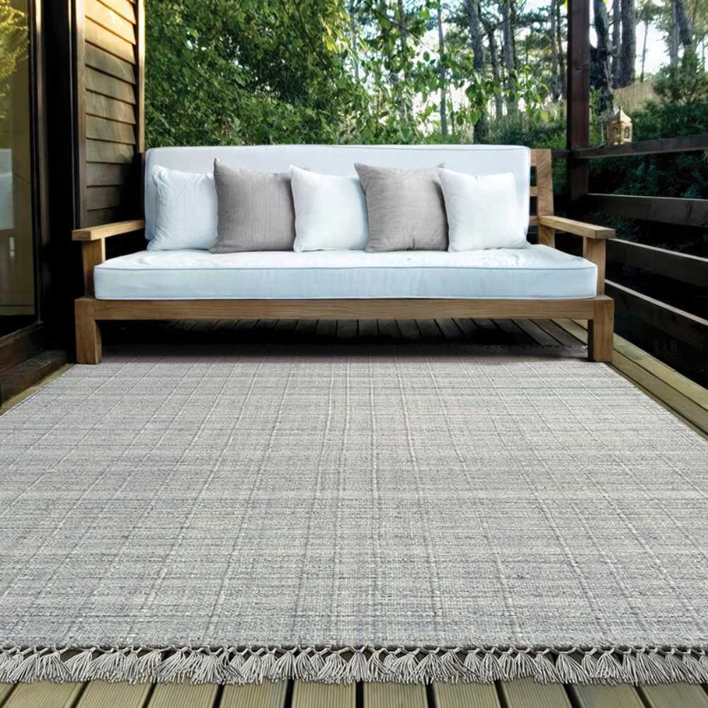 A full outdoor setting displays the Couristan Ascot Indoor/Outdoor Rug - Windowpane/Grey under a wooden bench with white cushions on a cozy deck