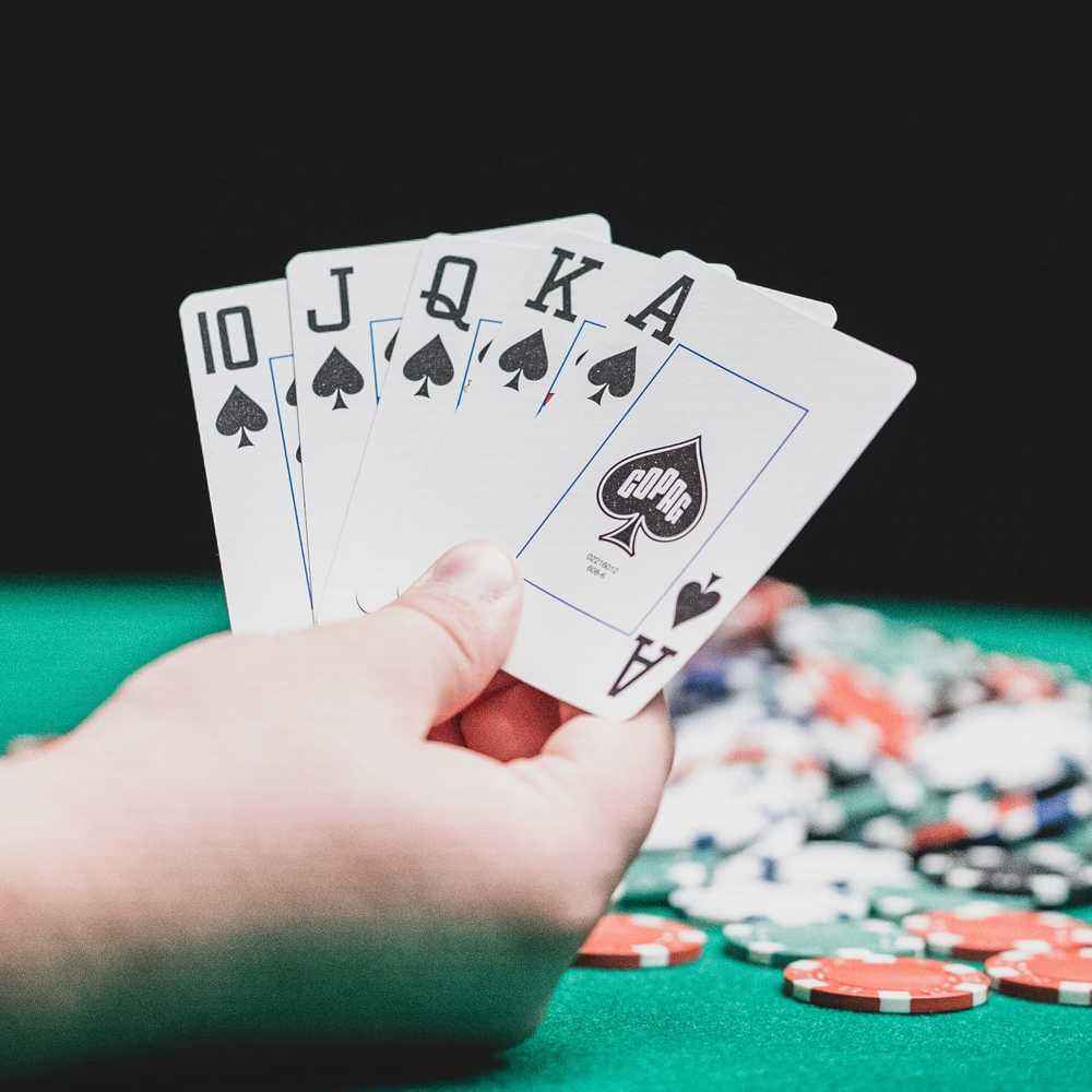 A hand holding a royal flush in spades using BBO Poker Tables Copag Masters 100% Plastic Playing Cards with poker chips blurred in the background