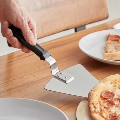 A hand uses the Chicago Brick Oven 13 14 Extra Large Pizza Pie Server with Black Offset Handle to lift a slice of pepperoni pizza from a wooden cutting board