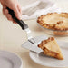 A hand uses the Chicago Brick Oven 13 14 Extra Large Pizza Pie Server with Black Offset Handle to serve a slice of apple pie onto a plate