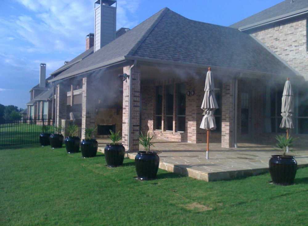 A large brick home patio with umbrellas and potted plants is cooled by the Aeromist Pinnacle 1000 PSI Misting System spraying mist along the eaves