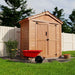 A left-angled view of the Outdoor Living Today SpaceMaster 6×4 with Double Door displays the shed’s side paneling and open yard space beside it, with a red wheelbarrow in front