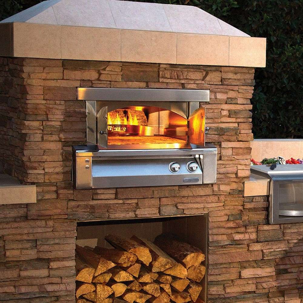 Alfresco Grills 30 Pizza Oven Plus - Built-In is shown installed in a stunning outdoor kitchen with a roaring fire inside, set in a rustic stone enclosure