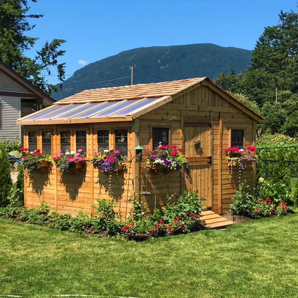 A lush backyard hosts an Outdoor Living Today 12×12 Sunshed Garden Shed lined with vibrant flower boxes and surrounded by a neatly landscaped garden