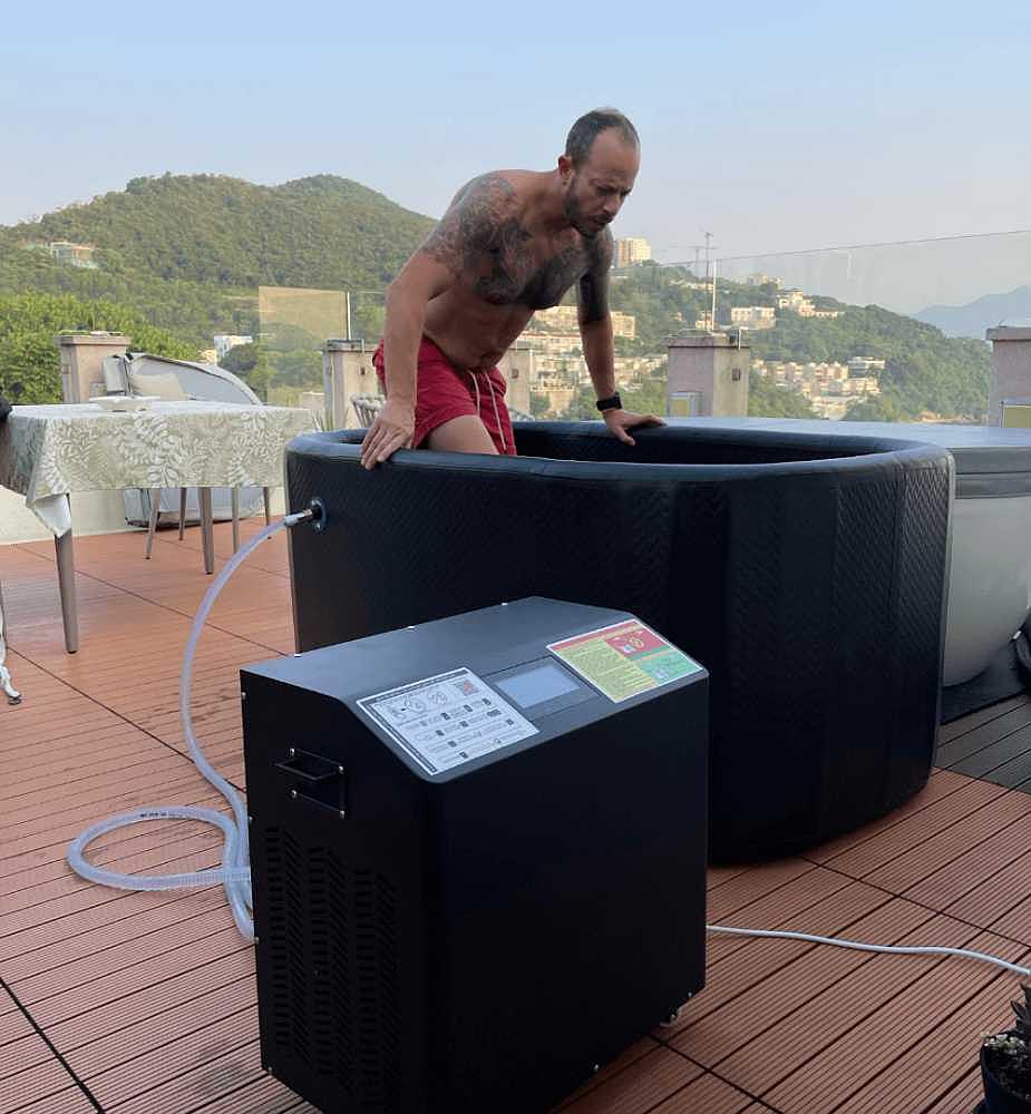 A man in red shorts is entering the Dreampod Ice Bath FLEX, connected to a black chiller unit on a sunlit rooftop deck