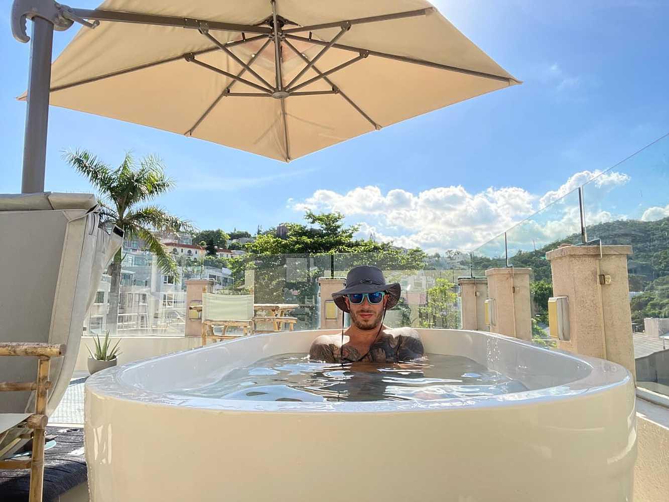 A man relaxes in a Dreampod Ice Bath with Chiller outdoors under a large umbrella with a tropical landscape in the background