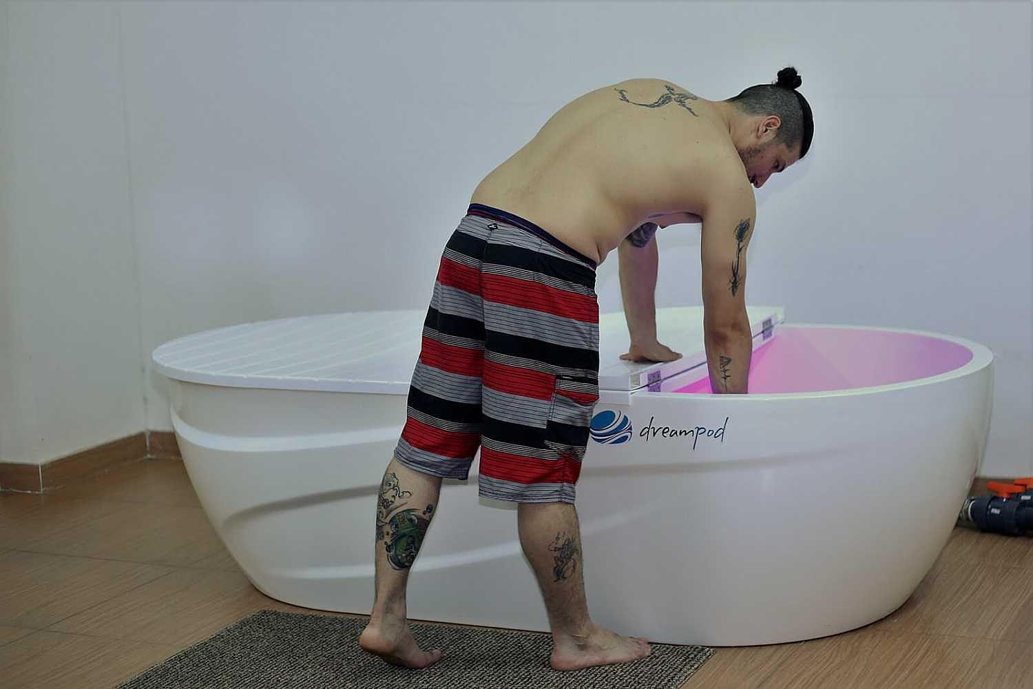 A man with tattoos leans over a Dreampod Home Float Pro, checking the water’s temperature inside the softly lit tank