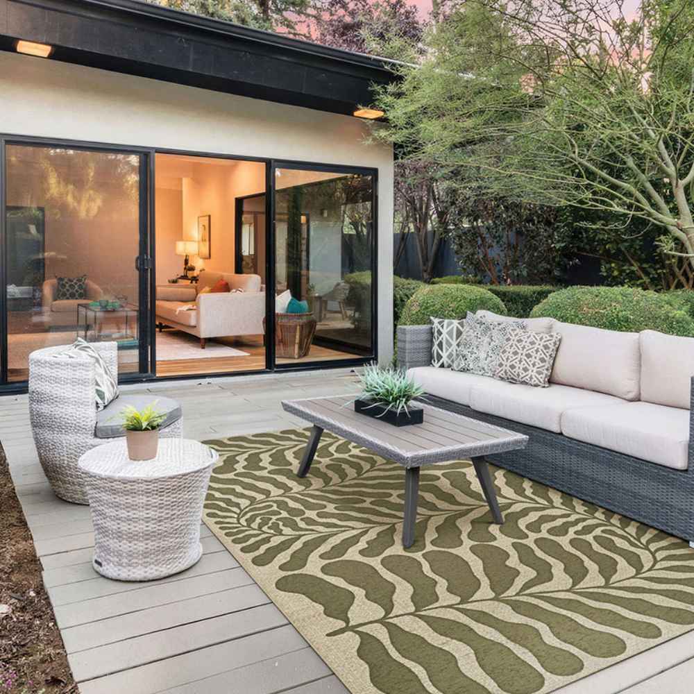 A modern patio setup features a cozy sofa and wicker furniture centered around the Couristan Castille Indoor/Outdoor Rug - Fiore/Tea Leaf